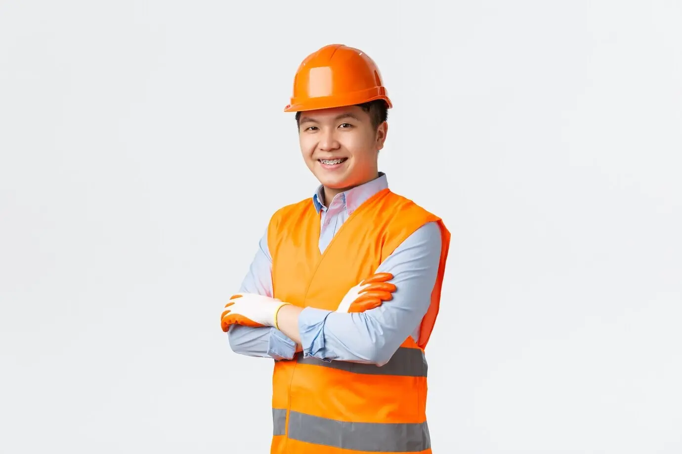 Konzept für Bauwesen und Industriearbeiter. Selbstbewusster junger asiatischer Ingenieur, Bauleiter, in reflektierender Kleidung und mit Helm, mit verschränkten Armen und frechem Lächeln, sorgt für Qualität, weiße Wand.
